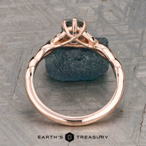 The "Kirryn" ring in 14K rose gold, featuring a 1.17-carat Montana sapphire and leaf motifs, is displayed upright on a rough blue stone background. Earth’s Treasury logo appears at the bottom.
