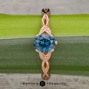 The "Kirryn" ring in 14K rose gold, set with a 1.17-carat Montana sapphire and leaf-inspired accents, stands upright on a green and gray surface with the Earth’s Treasury logo below.