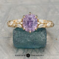 The gold cluster ring showcases an oval lavender gemstone centered between marquise and round diamonds, displayed on a textured blue-green stone.