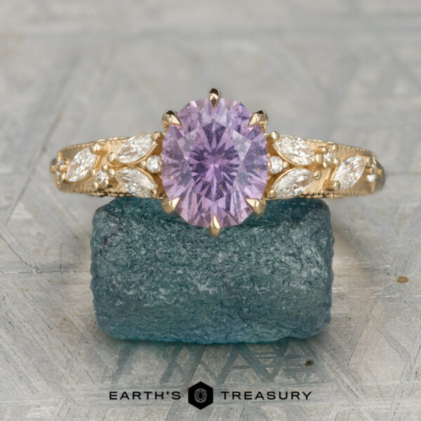 The "Chrysanthemum" ring in 14K yellow gold with 1.75-carat Umba sapphire The gold cluster ring showcases an oval lavender gemstone centered between marquise and round diamonds, displayed on a textured blue-green stone.