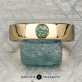 The "Logan" ring in 18K yellow gold, featuring an oval green gemstone, is displayed on a rough blue-green stone with the "EARTH'S TREASURY" logo beneath.