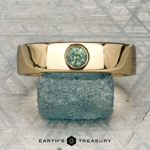 The "Logan" ring in 18K yellow gold, featuring an oval green gemstone, is displayed on a rough blue-green stone with the "EARTH'S TREASURY" logo beneath.