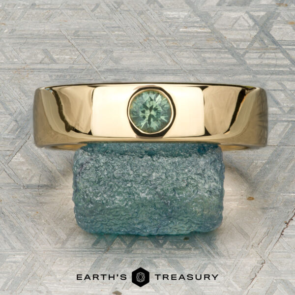 The "Logan" ring in 18K yellow gold The "Logan" ring in 18K yellow gold, featuring an oval green gemstone, is displayed on a rough blue-green stone with the "EARTH'S TREASURY" logo beneath.