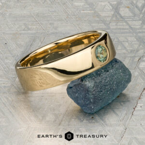 The "Logan" ring in 18K yellow gold, featuring an oval green gemstone, is displayed on a rough blue-green stone with the "EARTH'S TREASURY" logo beneath.