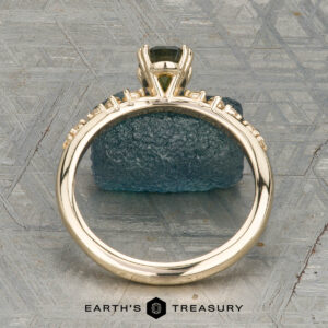 The "Blythe" ring in 14K yellow gold showcases a 0.90-carat Australian sapphire and diamond accents, displayed on a blue-green stone with the "Earth's Treasury" logo below.
