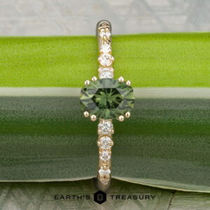 A 14K yellow gold "Blythe" ring with a 0.90-carat Australian sapphire and diamond accents on the band rests on a green leaf, with "EARTH'S TREASURY" at the bottom.