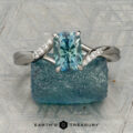 The "Tango" ring in platinum, featuring a 1.39-carat rectangular light blue Montana sapphire and diamond accents, is displayed upright on rough blue mineral with a patterned background and the "Earth's Treasury" logo below.