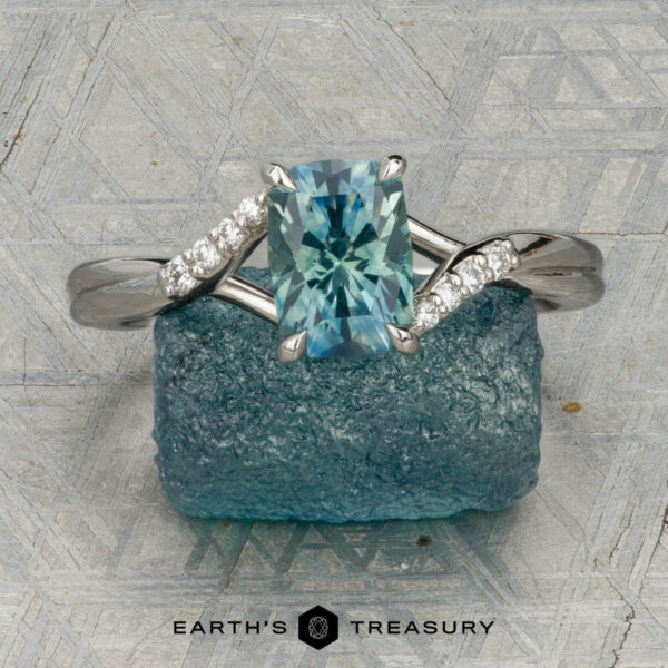 The "Tango" ring in platinum with 1.39-carat Montana sapphire The "Tango" ring in platinum, featuring a 1.39-carat rectangular light blue Montana sapphire and diamond accents, is displayed upright on rough blue mineral with a patterned background and the "Earth's Treasury" logo below.