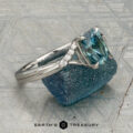 The "Tango" ring in platinum, featuring a 1.39-carat rectangular light blue Montana sapphire and diamond accents, is displayed upright on rough blue mineral with a patterned background and the "Earth's Treasury" logo below.