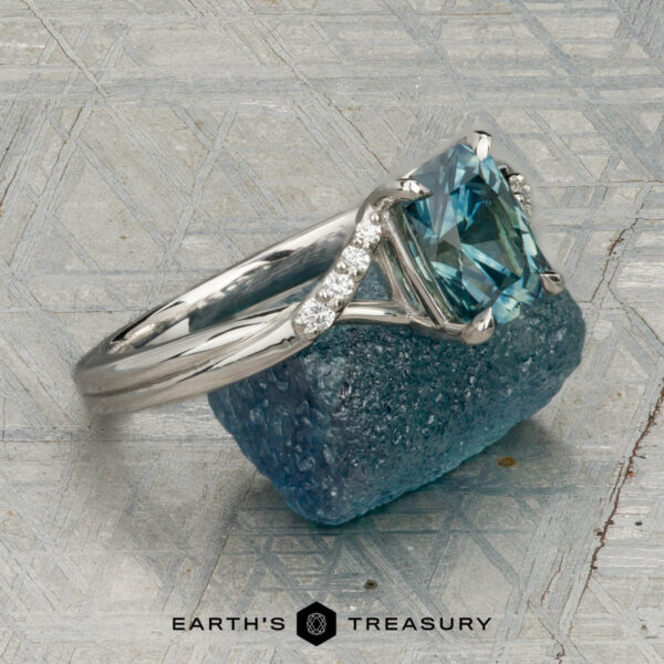 The "Tango" ring in platinum with 1.39-carat Montana sapphire The "Tango" ring in platinum, featuring a 1.39-carat rectangular light blue Montana sapphire and diamond accents, is displayed upright on rough blue mineral with a patterned background and the "Earth's Treasury" logo below.