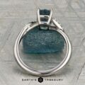 The "Tango" ring in platinum, featuring a 1.39-carat rectangular light blue Montana sapphire and diamond accents, is displayed upright on rough blue mineral with a patterned background and the "Earth's Treasury" logo below.