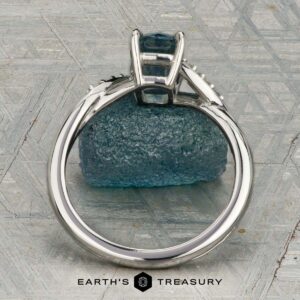 The "Tango" ring in platinum, featuring a 1.39-carat rectangular light blue Montana sapphire and diamond accents, is displayed upright on rough blue mineral with a patterned background and the "Earth's Treasury" logo below.