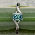 The "Tango" ring in platinum showcases a 1.39-carat rectangular Montana sapphire at its center with diamond accents on a curved band, displayed on a green plant stem.