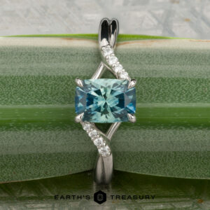 The "Tango" ring in platinum showcases a 1.39-carat rectangular Montana sapphire at its center with diamond accents on a curved band, displayed on a green plant stem.