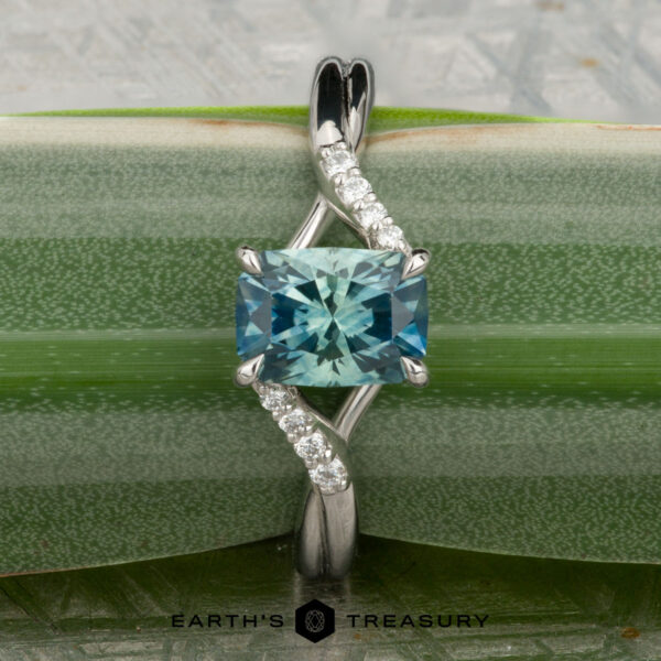 The "Tango" ring in platinum showcases a 1.39-carat rectangular Montana sapphire at its center with diamond accents on a curved band, displayed on a green plant stem.