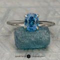 The silver solitaire ring with an oval blue gemstone stands upright on a rough blue stone, with the Earth's Treasury logo below.