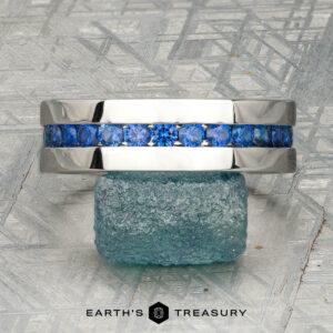 A platinum Atlas ring encircling a rough blue gemstone, displayed on a textured gray surface with "Earth's Treasury" at the bottom.