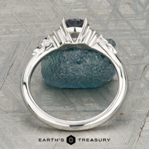The "Aster" ring in platinum, featuring a 0.80-carat Montana sapphire and accent stones, rests on rough blue crystal with textured geometric patterns in the background.
