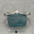 The "Aster" band in platinum, featuring a leaf motif and central diamond, is displayed atop a rough blue gemstone on a patterned surface with the text "EARTH'S TREASURY" below.