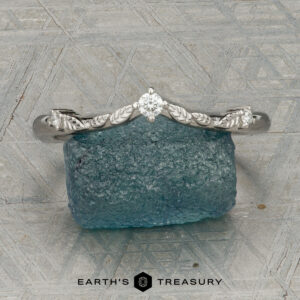 The "Aster" band in platinum, featuring a leaf motif and central diamond, is displayed atop a rough blue gemstone on a patterned surface with the text "EARTH'S TREASURY" below.