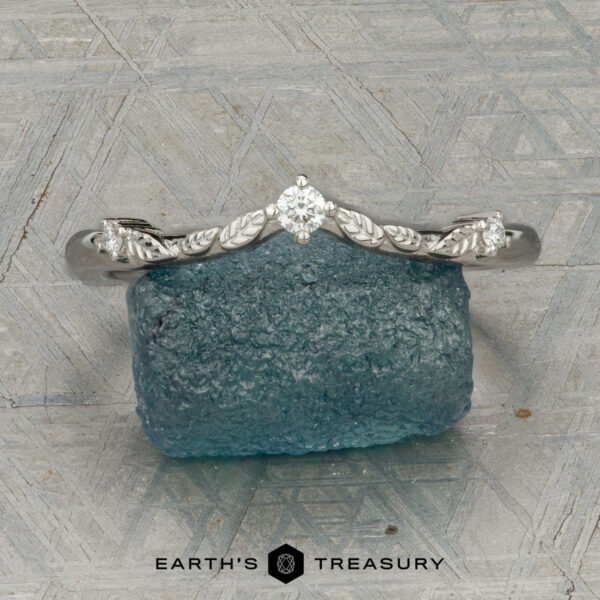 The "Aster" band in platinum The "Aster" band in platinum, featuring a leaf motif and central diamond, is displayed atop a rough blue gemstone on a patterned surface with the text "EARTH'S TREASURY" below.