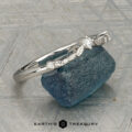 The "Aster" band in platinum, featuring a leaf motif and central diamond, is displayed atop a rough blue gemstone on a patterned surface with the text "EARTH'S TREASURY" below.