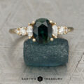 The "Tigrina" ring in 14K yellow gold features a 1.94-carat oval green Australian sapphire, two marquise and two round side diamonds, displayed on a rough blue stone with the "Earth's Treasury" logo at the bottom.