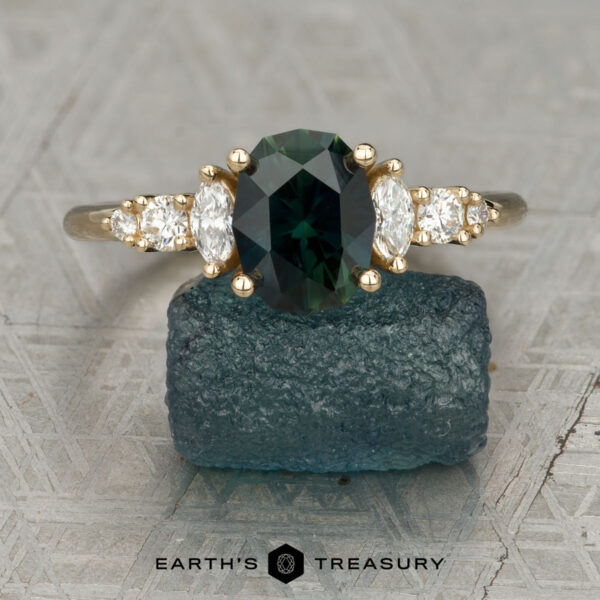 The "Tigrina" ring in 14K yellow gold with 1.94-carat Australian sapphire The "Tigrina" ring in 14K yellow gold features a 1.94-carat oval green Australian sapphire, two marquise and two round side diamonds, displayed on a rough blue stone with the "Earth's Treasury" logo at the bottom.