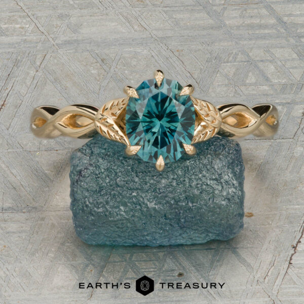 The "Kirryn" ring in 14K yellow gold with 1.37-carat Montana sapphire The "Kirryn" ring in 14K yellow gold with a 1.37-carat Montana sapphire, featuring leaf accents, is shown on a rough blue mineral with the "Earth's Treasury" logo below.