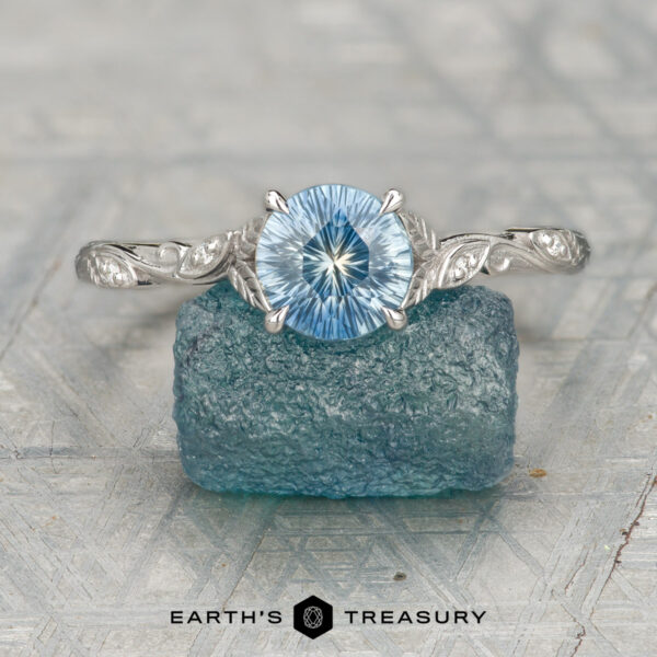 Amborella Ring The Amborella ring showcases a round blue gemstone that rests elegantly atop a rough blue crystal. The textured grey background adds depth to the scene, with the "Earth's Treasury" logo and text featured at the bottom of the image.