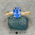 The gold ring with an oval blue gemstone and diamond-accented solitaire design is displayed on a rough blue-green stone, with the Earth's Treasury logo below.