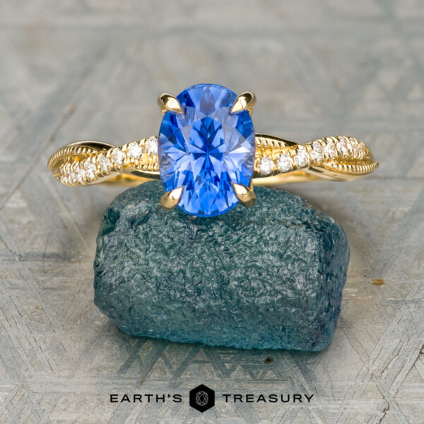 The "Musette" ring in 18K yellow gold with 1.88-carat Ceylon sapphire The gold ring with an oval blue gemstone and diamond-accented solitaire design is displayed on a rough blue-green stone, with the Earth's Treasury logo below.