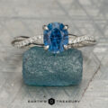The silver solitaire ring features a blue oval gemstone and clear diamond accents on the band, displayed atop a rough blue crystal against a light gray geometric background.