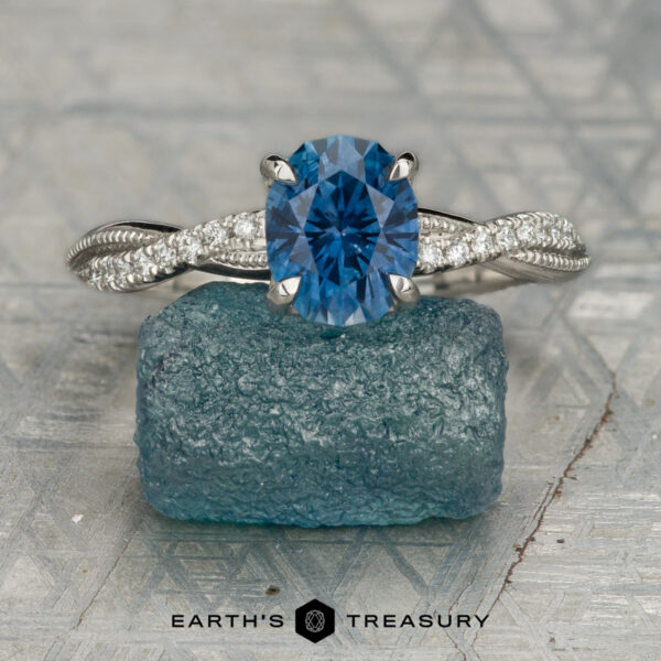 The "Musette" ring in platinum with 1.53-carat Montana sapphire The silver solitaire ring features a blue oval gemstone and clear diamond accents on the band, displayed atop a rough blue crystal against a light gray geometric background.