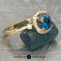 The “Eleanor” ring in 18K yellow gold, featuring a 1.47-carat Montana sapphire and diamond halo, is displayed on rough blue mineral with the Earth’s Treasury logo below.