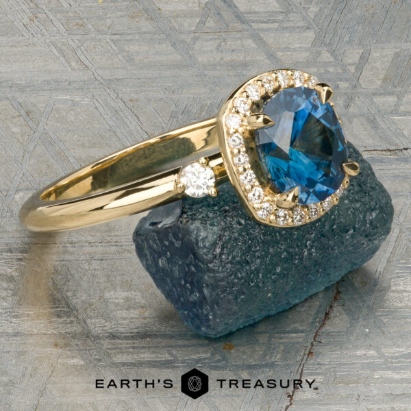 The “Eleanor” ring in 18K yellow gold with 1.47-carat Montana sapphire The “Eleanor” ring in 18K yellow gold, featuring a 1.47-carat Montana sapphire and diamond halo, is displayed on rough blue mineral with the Earth’s Treasury logo below.