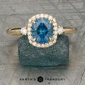 The “Eleanor” ring in 18K yellow gold, featuring a 1.47-carat Montana sapphire and diamond halo, is displayed on rough blue mineral with the Earth’s Treasury logo below.