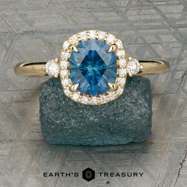 The “Eleanor” ring in 18K yellow gold with 1.47-carat Montana sapphire The “Eleanor” ring in 18K yellow gold, featuring a 1.47-carat Montana sapphire and diamond halo, is displayed on rough blue mineral with the Earth’s Treasury logo below.