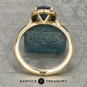 The “Eleanor” ring in 18K yellow gold, featuring a 1.47-carat Montana sapphire and diamond halo, is displayed on rough blue mineral with the Earth’s Treasury logo below.