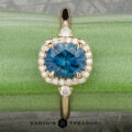 The “Eleanor” ring in 18K yellow gold, featuring a 1.47-carat Montana sapphire and diamond halo, is displayed on rough blue mineral with the Earth’s Treasury logo below.