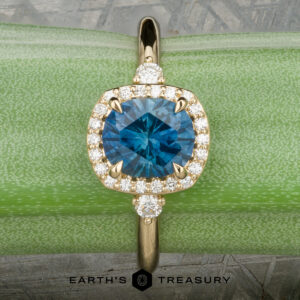 The “Eleanor” ring in 18K yellow gold, featuring a 1.47-carat Montana sapphire and diamond halo, is displayed on rough blue mineral with the Earth’s Treasury logo below.