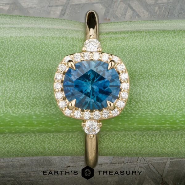 The “Eleanor” ring in 18K yellow gold with 1.47-carat Montana sapphire The “Eleanor” ring in 18K yellow gold, featuring a 1.47-carat Montana sapphire and diamond halo, is displayed on rough blue mineral with the Earth’s Treasury logo below.