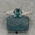 A blue-green "Stacked from 14 images. Method=B (R=14,S=2)" ring with a detailed silver band rests on a rough blue stone, displayed on a textured surface. The "Earth's Treasury" logo is visible at the bottom.