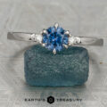 The Samia ring featuring a round blue gemstone and two small diamonds on each side, displayed upright on rough blue stone with the "Earth's Treasury" logo below—an elegant three-stone design.