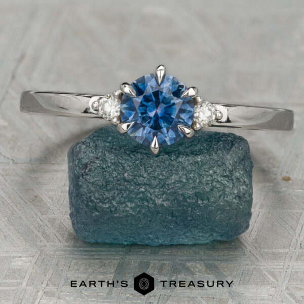 The Samia Ring The Samia ring featuring a round blue gemstone and two small diamonds on each side, displayed upright on rough blue stone with the "Earth's Treasury" logo below—an elegant three-stone design.
