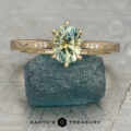 The Carmen ring featuring an oval light green gemstone with small diamonds on the band rests on a blue-green stone. The "EARTH'S TREASURY" logo and text are visible below.