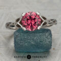 A silver solitaire ring featuring a round pink gemstone rests on a rough blue stone. The Earth's Treasury logo appears at the bottom of the image, drawing attention to the elegance of the Harper, Taylor design.