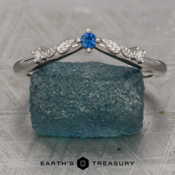 The "Aster" band in platinum A platinum "Aster" band with a blue center gemstone and two small clear stones rests on a rough blue crystal; "EARTH'S TREASURY" is written below.