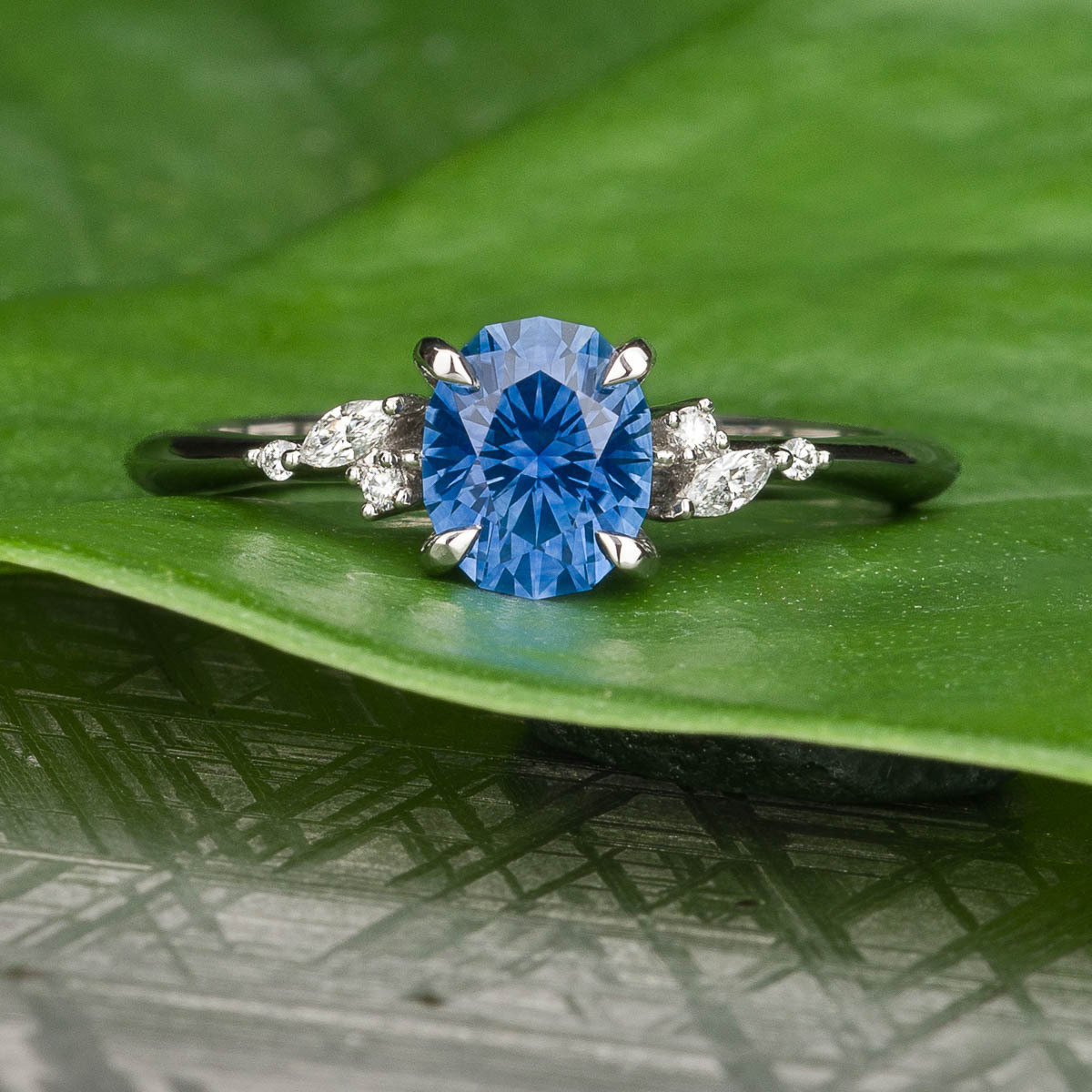 The All-Rings features a silver band with an oval blue gemstone center and two marquise-cut white stones, showcased on a green leaf background.
