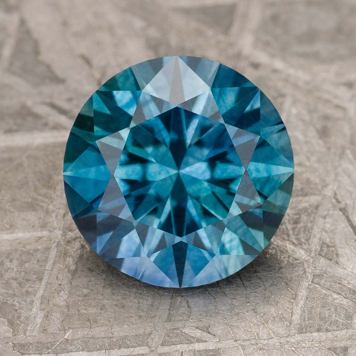A 2.56-carat rich teal Montana sapphire (heated), round brilliant-cut, rests on a textured metallic surface.